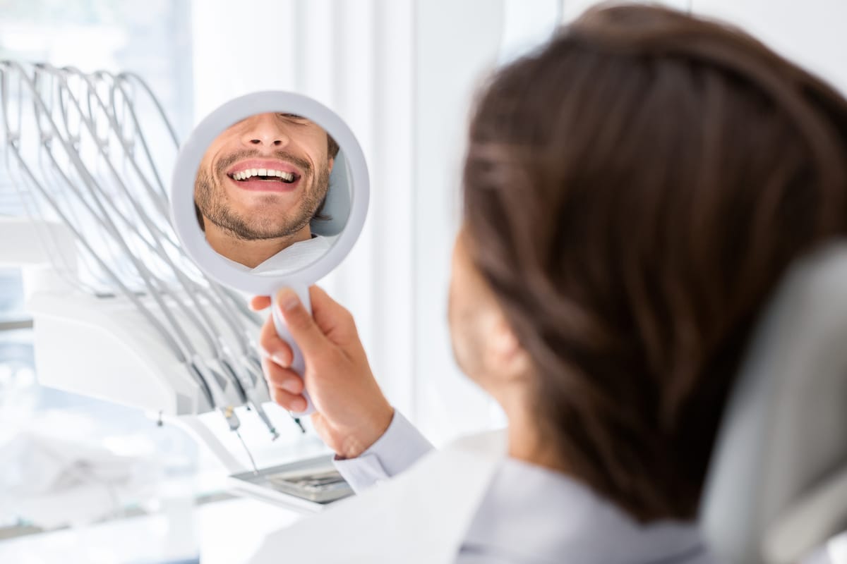 Satisfied man looking at mirror and checking his teeth in dental clinic, beautiful smile concept, cropped
